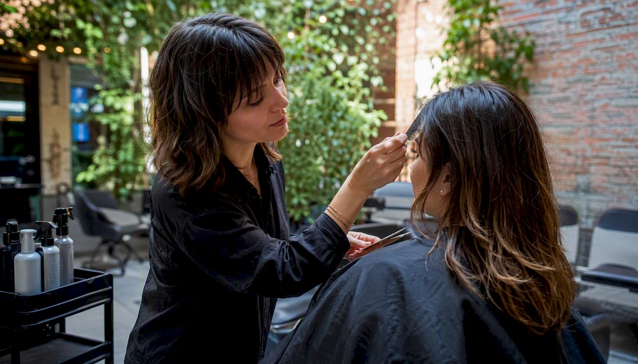 Makeup artist applying eye makeup to client outdoors, with greenery and brick wall in the background.